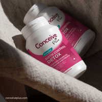 Two white Conceive Plus Birth Control Detox supplement bottles with pink labels, nestled in a beige linen fabric under warm natural sunlight.