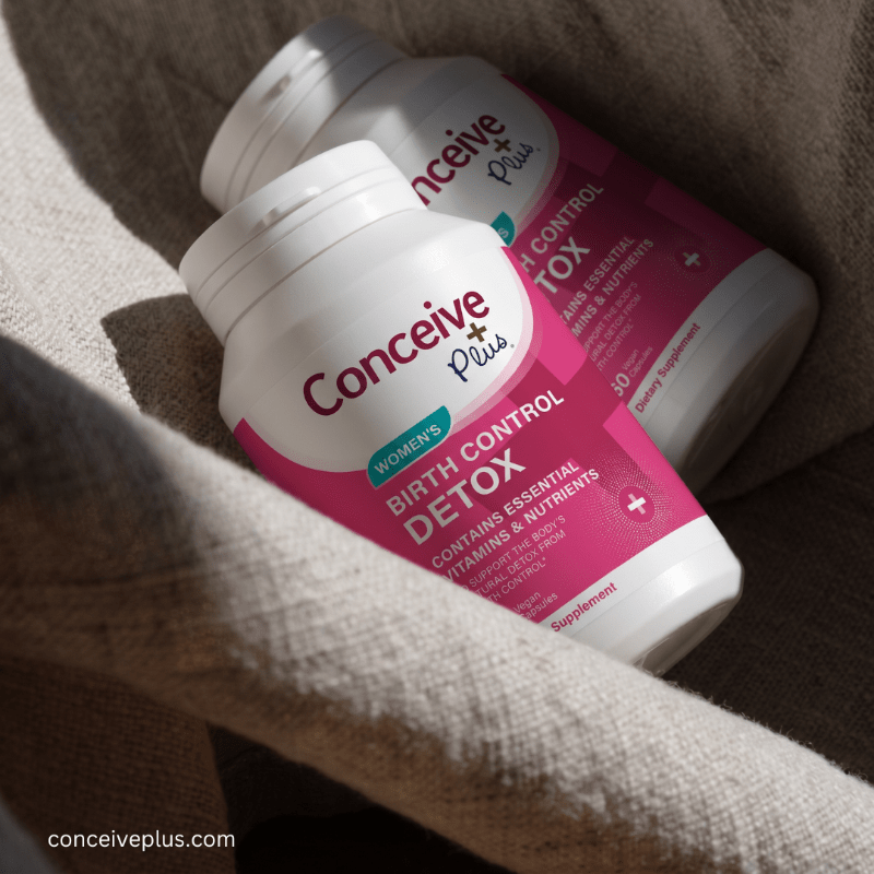 Two white Conceive Plus Birth Control Detox supplement bottles with pink labels, nestled in a beige linen fabric under warm natural sunlight.