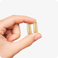 Close-up of a hand holding two beige Conceive Plus Prenatal capsules between the thumb and index finger, showcasing capsule size and texture against a plain white background.
