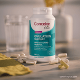 A bottle of Conceive Plus Women's Ovulation Support dietary supplement capsules sits on a wooden surface with scattered capsules and a soft-focus glass jar in the background.
