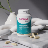 A bottle of Conceive Plus Prenatal food supplement stands on a white marble slab with capsules scattered around it. The bottle has a white cap and a teal and white label with pink and blue text.